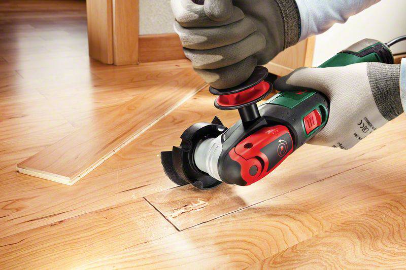 Une personne découpe un morceau rectangulaire dans un plancher en bois à l'aide d'un outil électrique portable. Des copeaux de bois sont visibles.