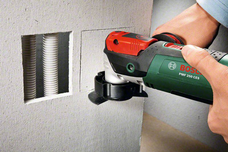 Une personne utilise un outil électrique vert pour découper une ouverture dans un mur. Un conduit de câbles est visible à droite.