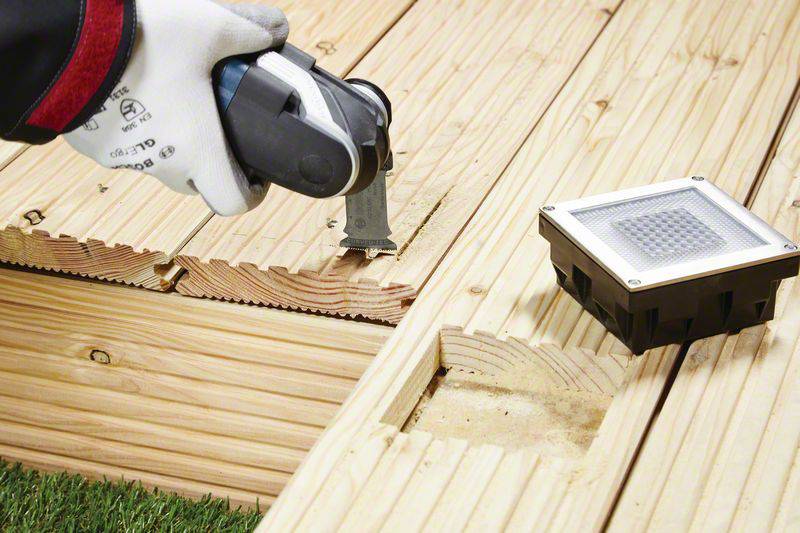 Une personne installe des panneaux en bois avec un outil électrique sur un plancher de terrasse. À côté se trouve une lampe de sol rectangulaire.