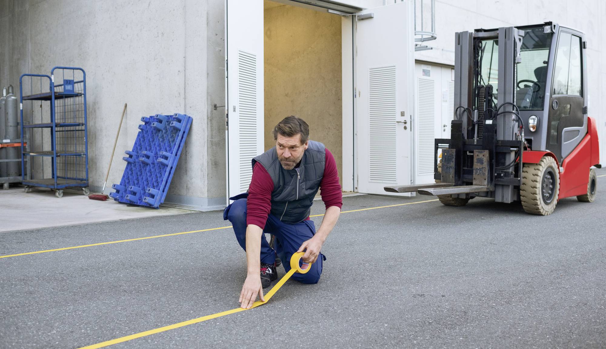 Un homme est à genoux sur le sol et colle du ruban de signalisation jaune sur l'asphalte devant un entrepôt. Un chariot élévateur est visible en arrière-plan.