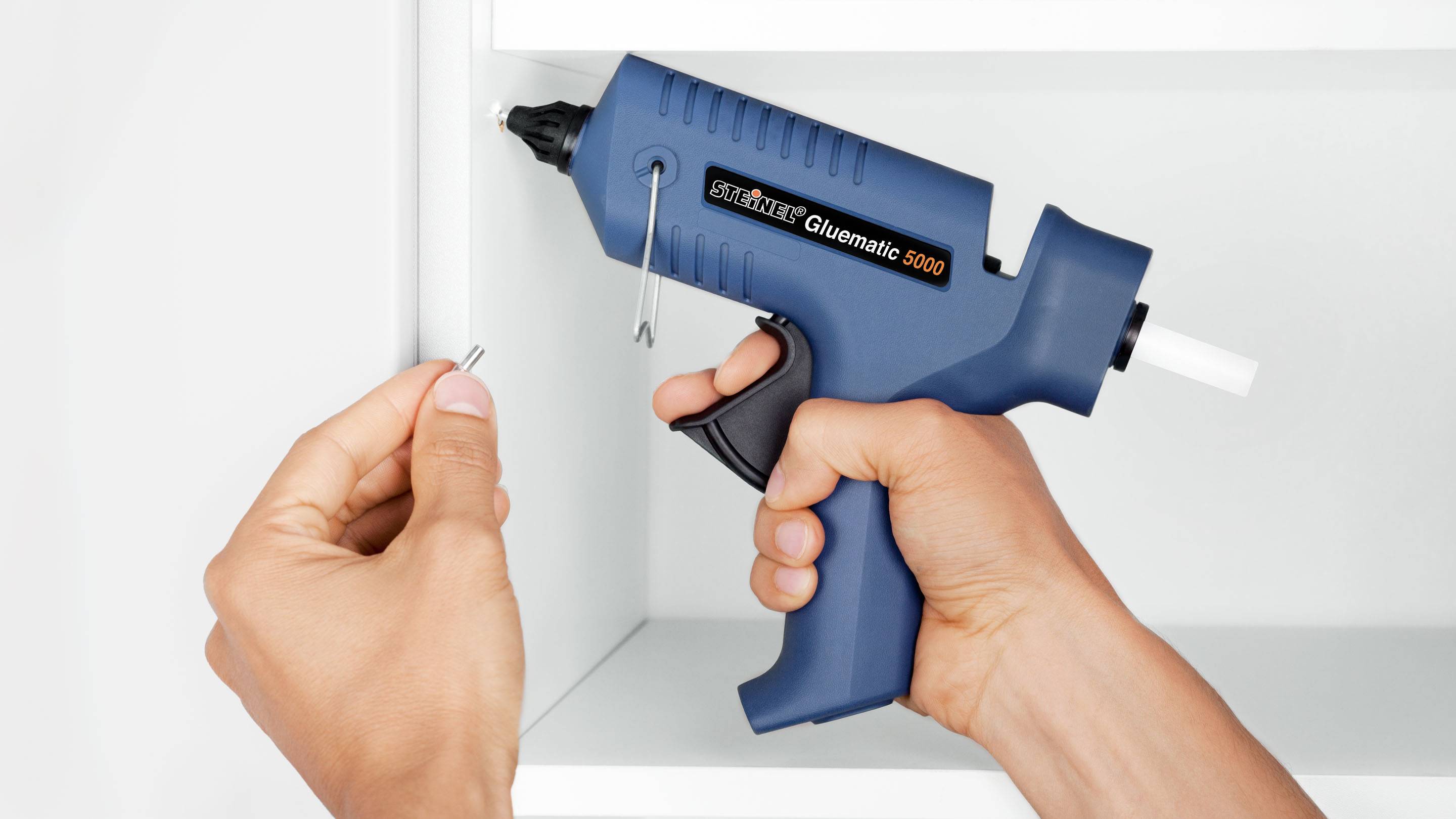 Une personne tient un pistolet à colle chaude 'Glunamatic 5000' et un petit clou devant une étagère blanche.