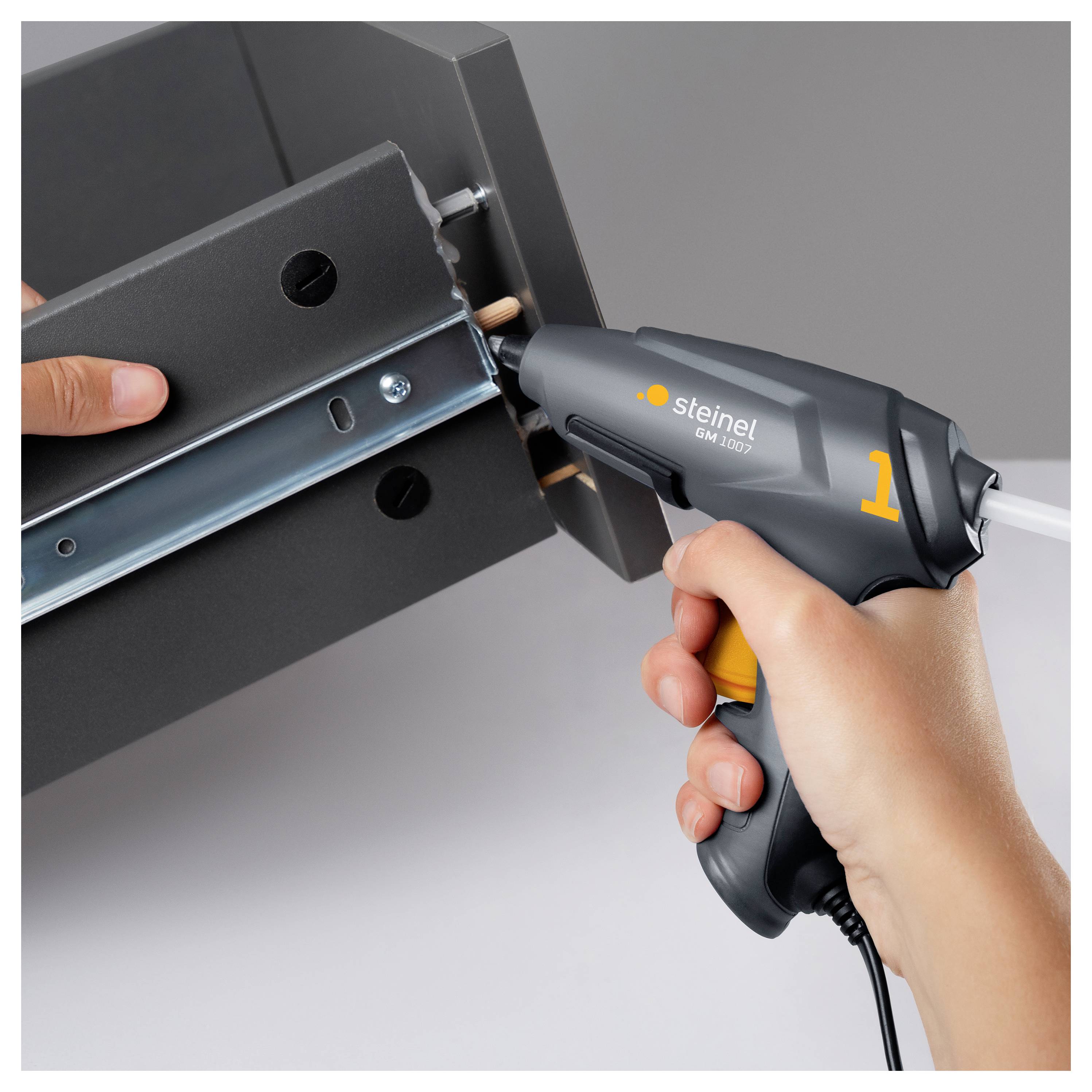 Des mains utilisant un pistolet thermique Steinel pour appliquer de la chaleur à un support métallique monté sur un panneau noir, probablement pour l'assemblage ou le retrait.
