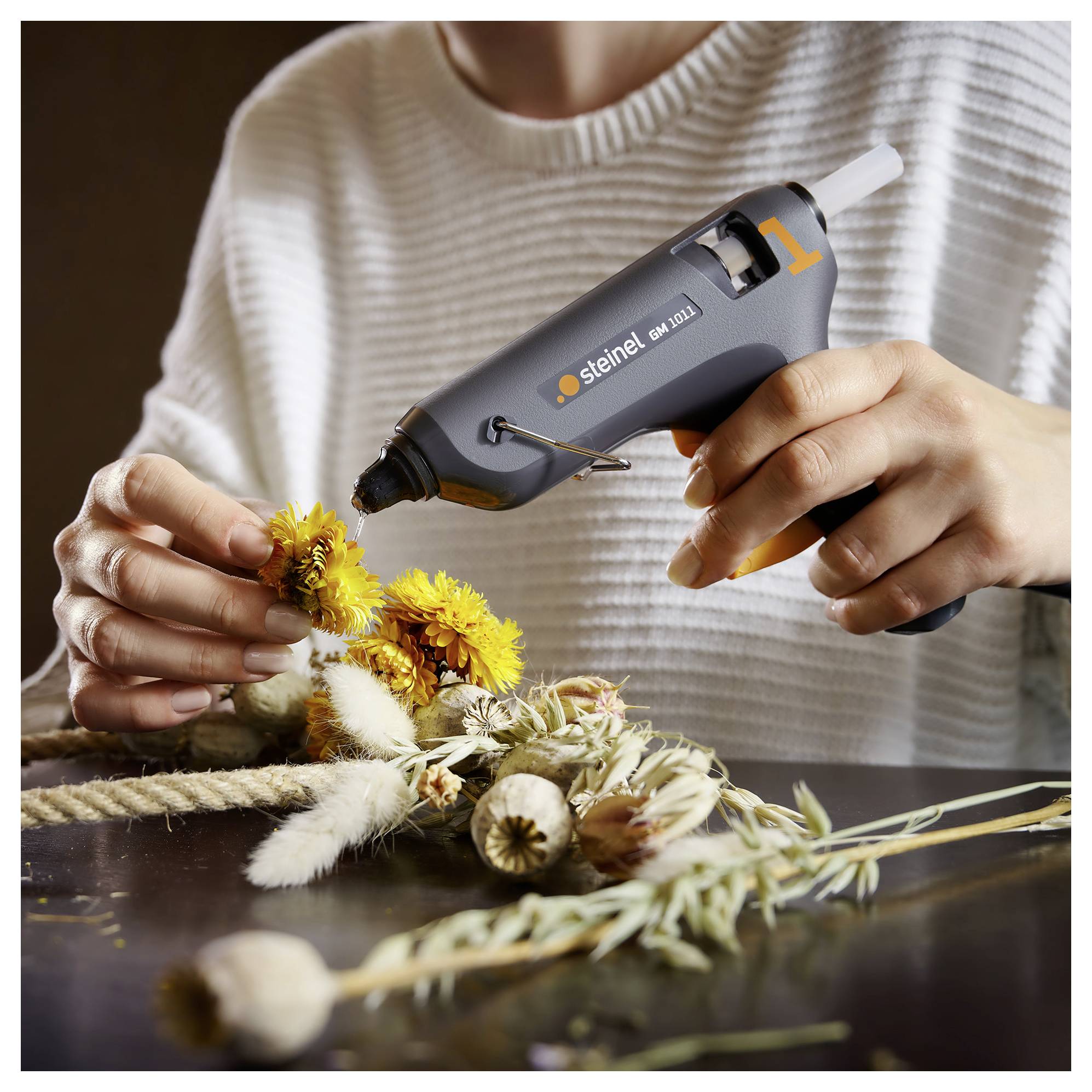 Une personne utilise un pistolet à colle chaude pour fixer des fleurs séchées à un arrangement artisanal sur une table.
