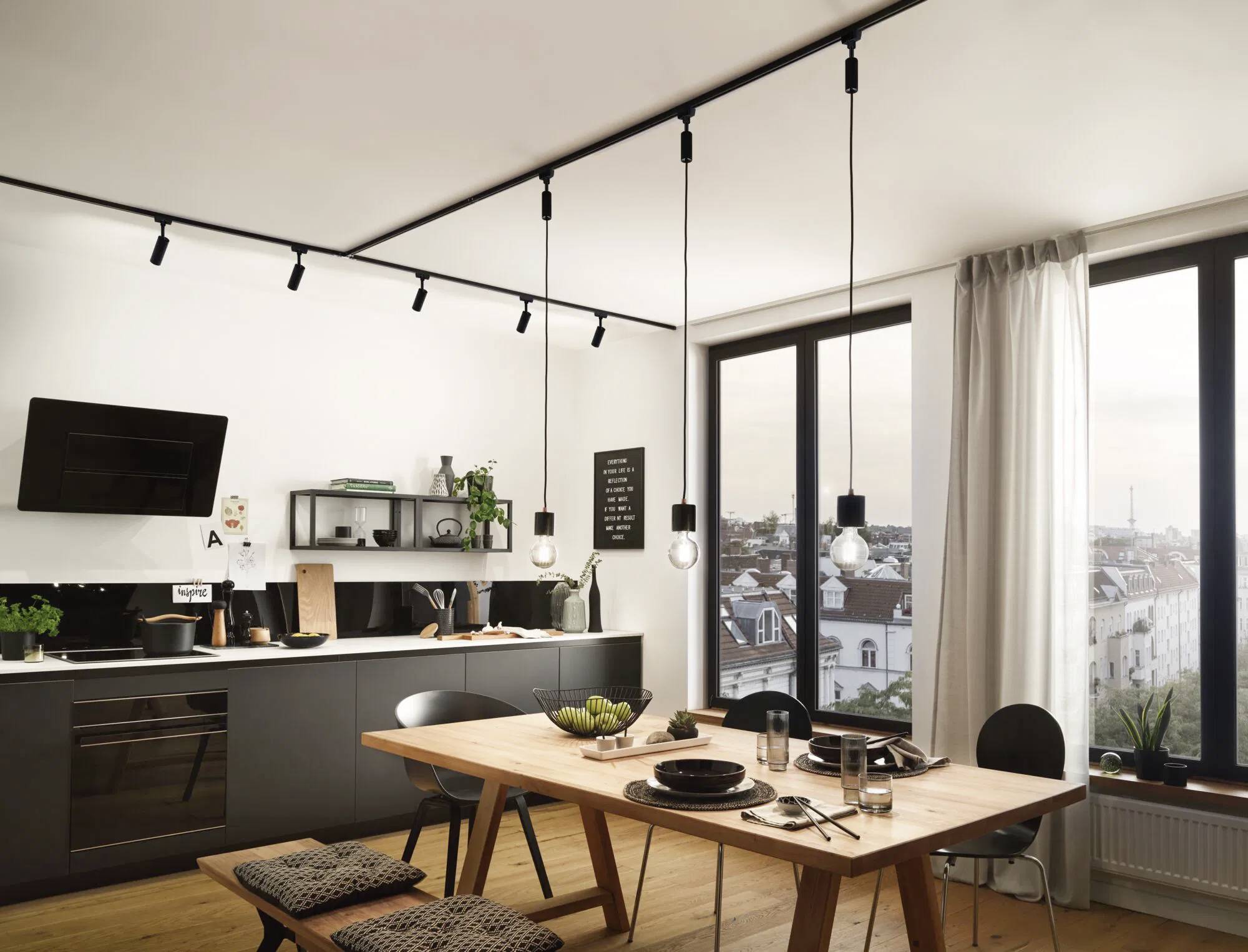 Cuisine moderne avec espace repas, table en bois et chaises rembourrées. De grandes fenêtres offrent une vue sur la ville. Éclairage assuré par des suspensions.