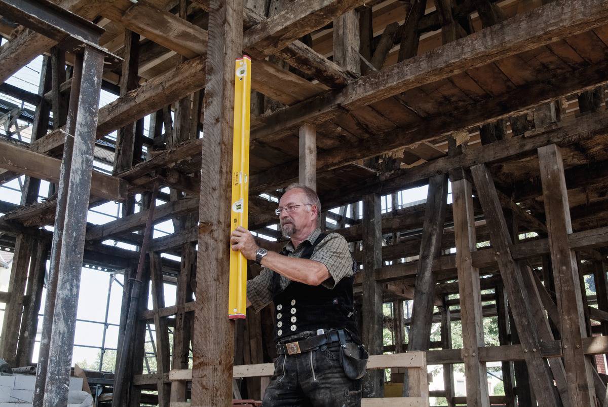 Un homme vêtu de vêtements de travail utilise un niveau à bulle pour mesurer un cadre en bois dans un bâtiment en bois inachevé.