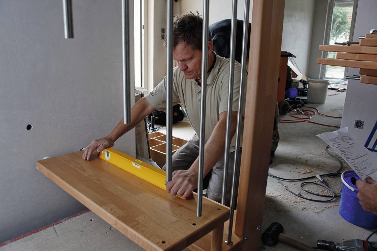 Un homme utilise un niveau à bulle pour installer une étagère en bois dans une maison. Des outils de construction sont visibles en arrière-plan.