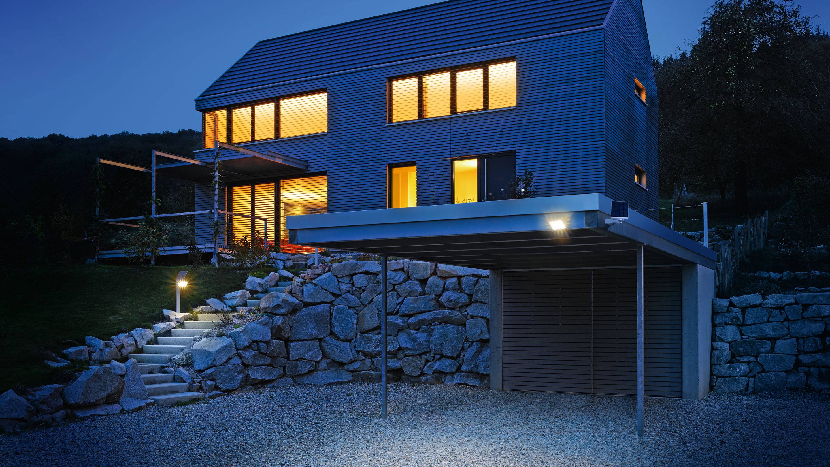 Maison moderne avec de grandes fenêtres et un éclairage de façade bleu la nuit, avec un escalier en pierre et un carport à l'avant-plan.