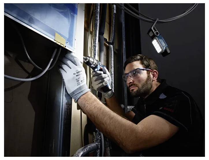 Un homme portant des lunettes de sécurité et des gants utilise une perceuse électrique pour travailler sur une surface métallique. Il est concentré et entouré de tuyaux.