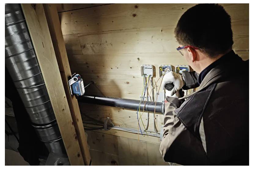 Une personne travaille sur le câblage électrique à l'intérieur d'une structure en bois, entourée de conduits et de tuyaux isolés. Un équipement de protection est porté.