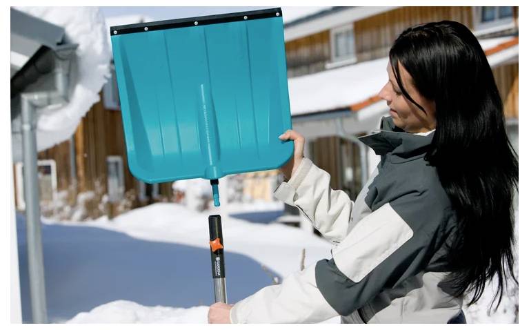 Une personne vêtue de vêtements d'hiver tient une pelle à neige à l'extérieur, avec des bâtiments couverts de neige visibles en arrière-plan.