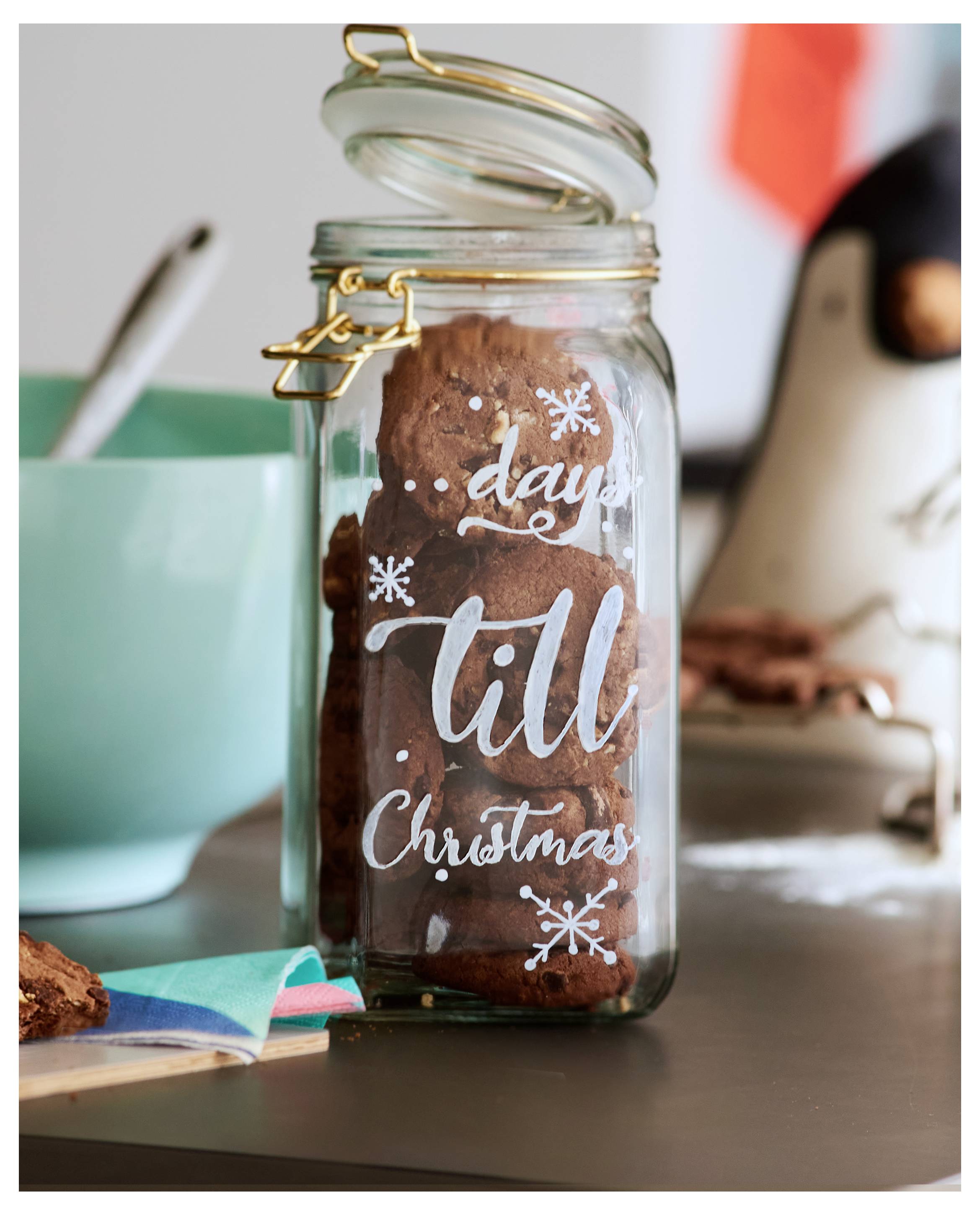 Bocal en verre rempli de biscuits au chocolat, étiqueté « jours jusqu'à Noël », sur une table avec un bol et des ustensiles en arrière-plan.