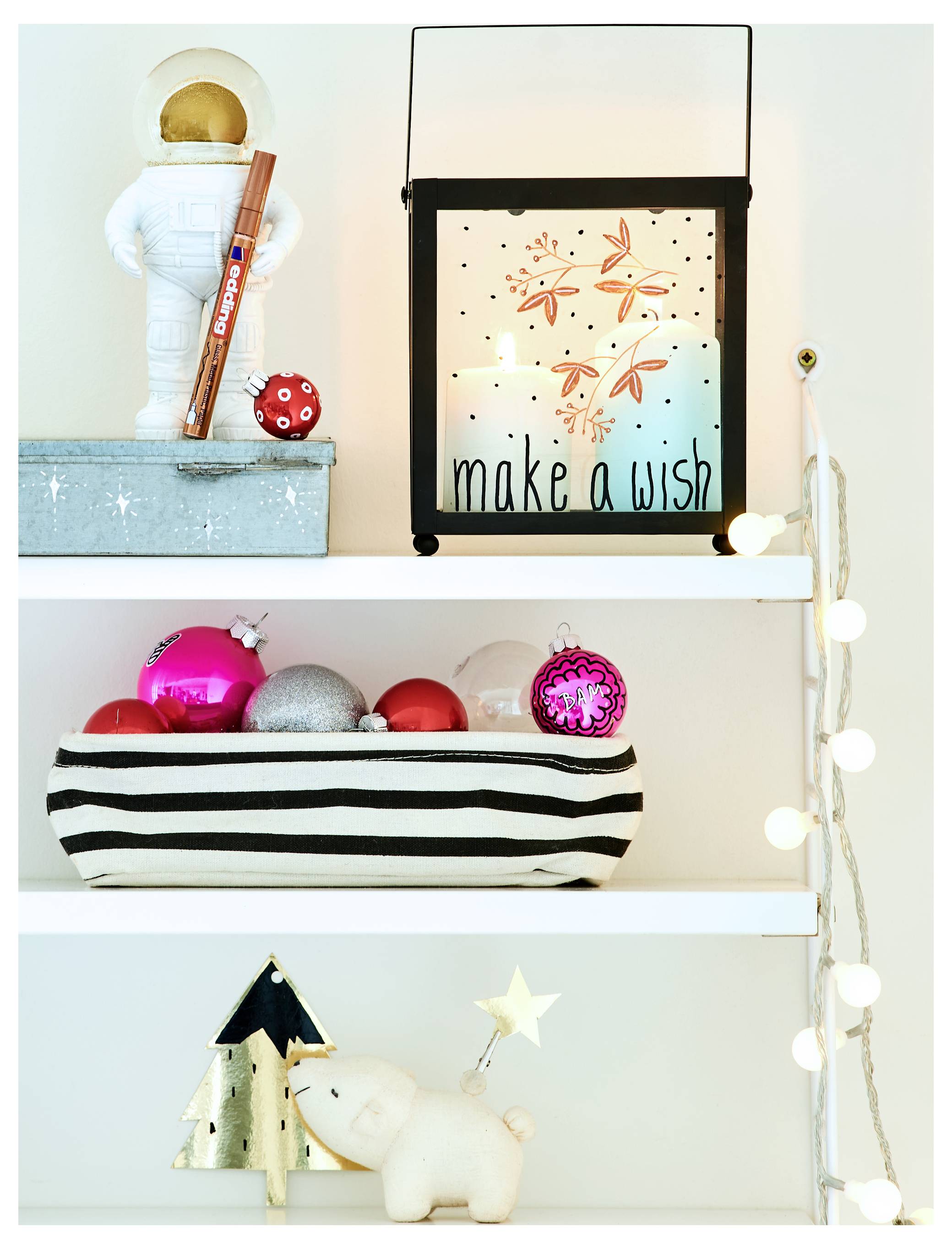 Une unité de rangement blanche avec des articles de décoration pour les vacances, notamment une lanterne « faire un vœu », un bol rayé rempli de boules de Noël, et de petites figurines d'arbre et de cochon avec des guirlandes lumineuses.