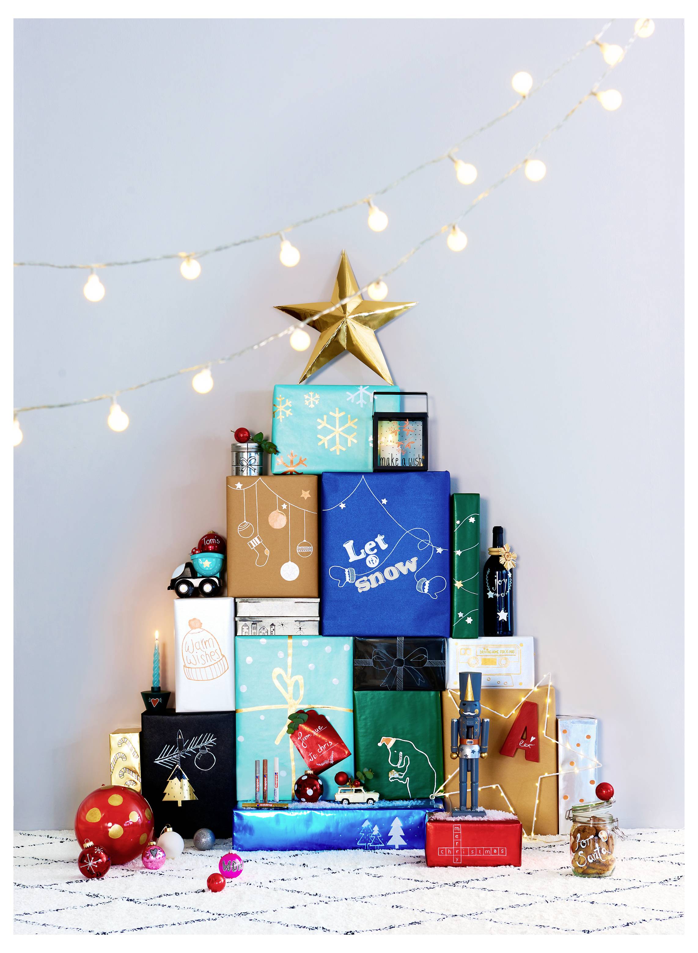 Pile festive de cadeaux emballés et de décoration de vacances sous des guirlandes lumineuses, surmontée d'une étoile dorée contre un mur pâle.