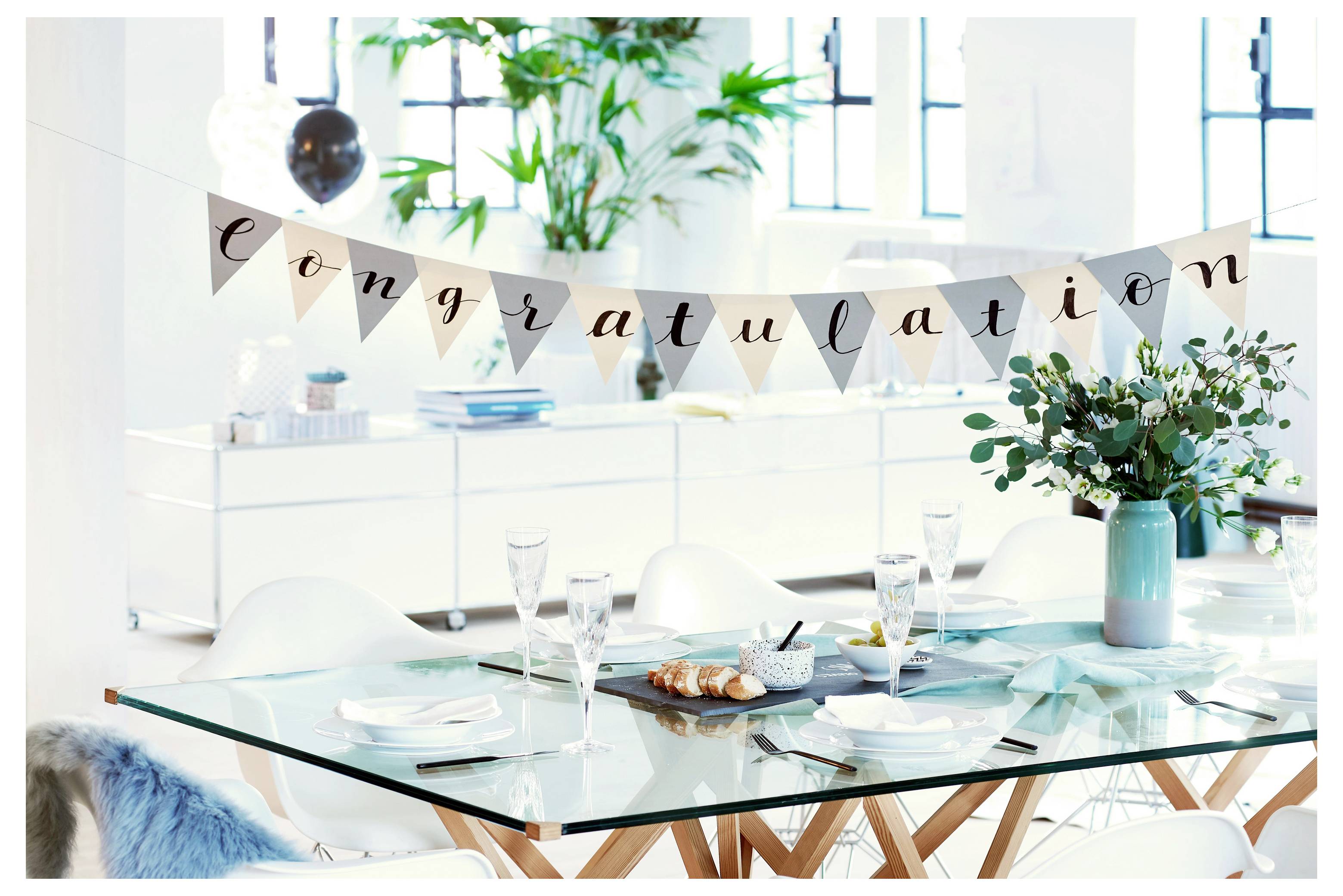 Salle à manger lumineuse décorée pour une célébration, avec une bannière « félicitations » au-dessus d'une table dressée et des fleurs dans un vase.