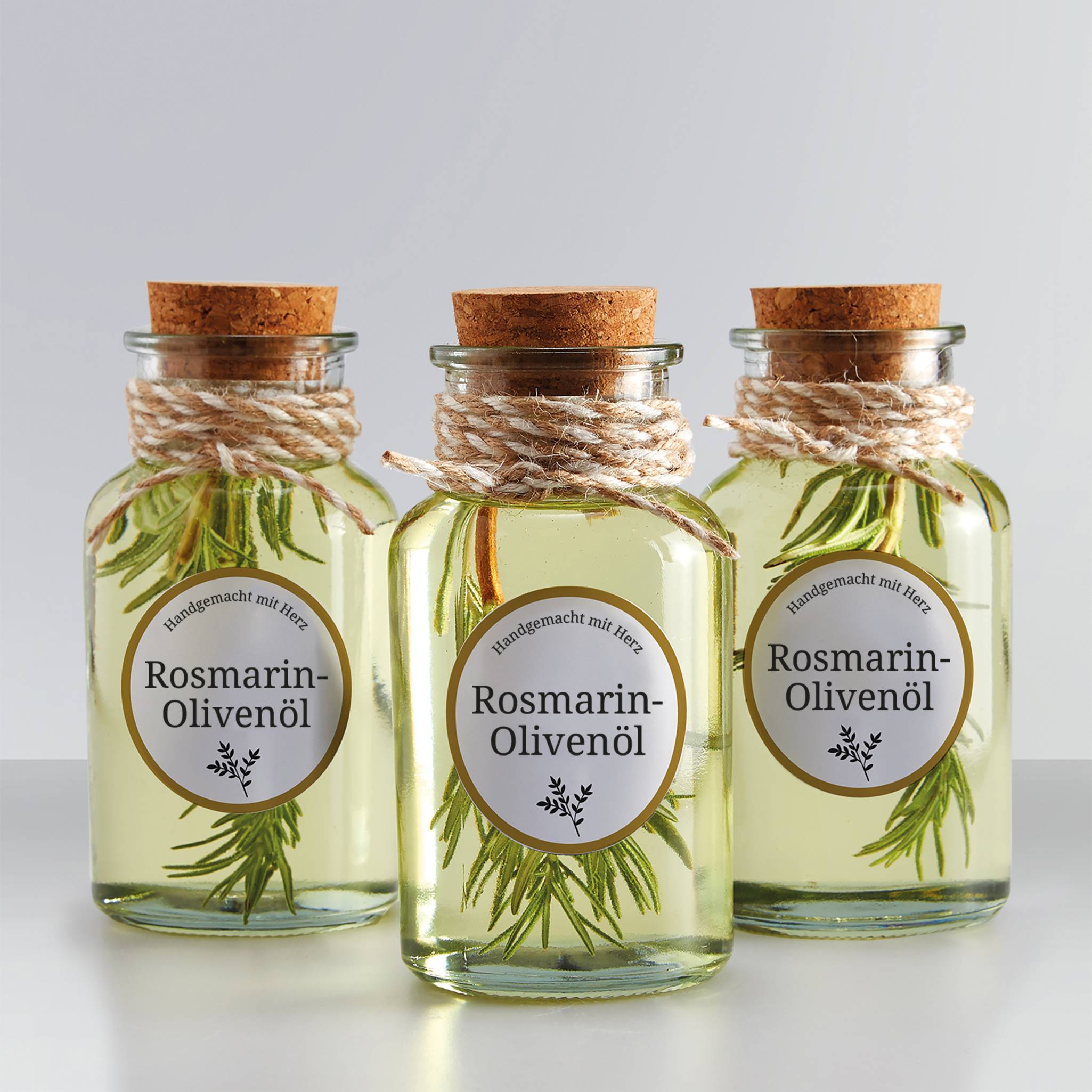Trois bouteilles en verre avec bouchons, remplies d'huile d'olive au romarin, décorées avec des branches de romarin, étiquetées 'Huile d'olive au romarin'.