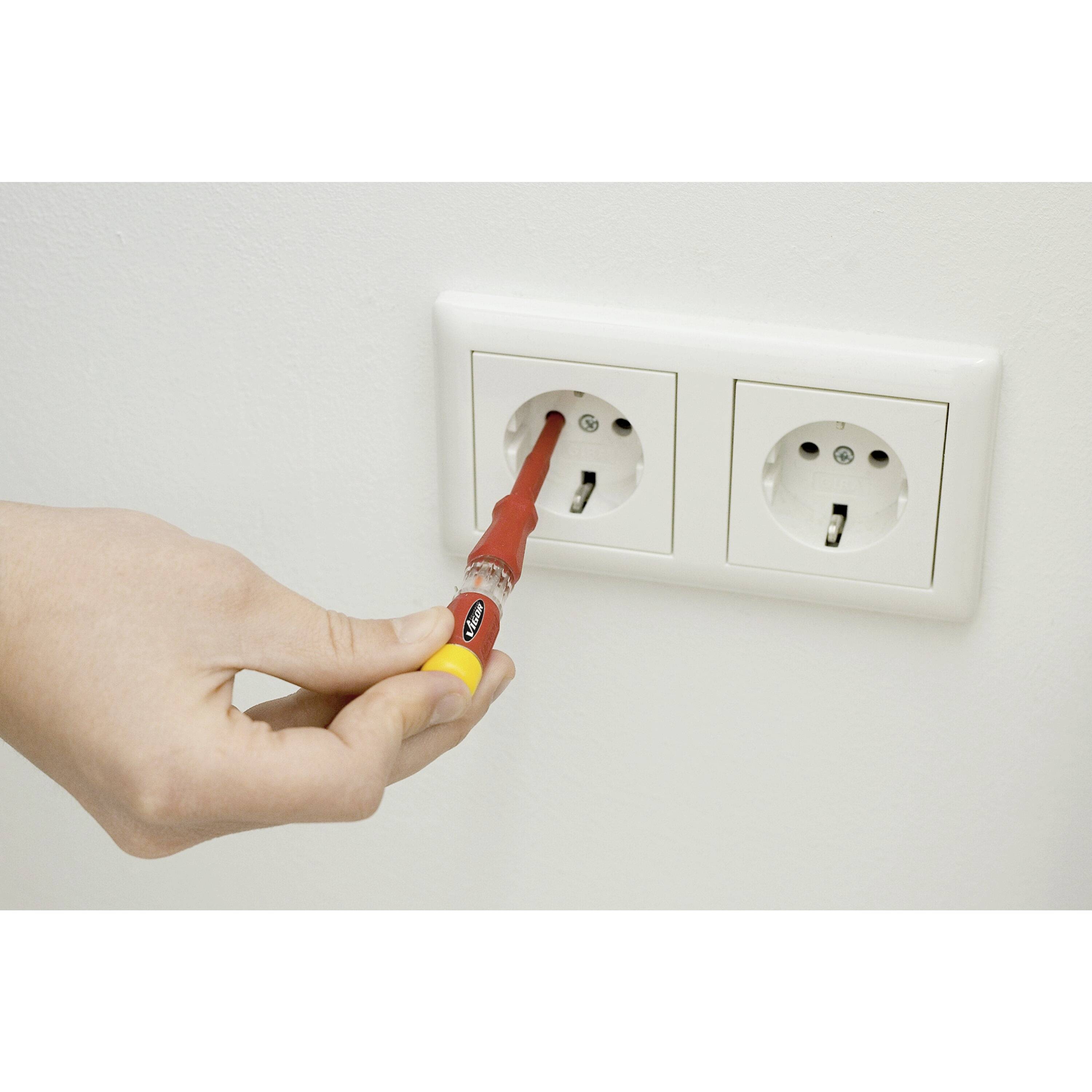 Une main tient un testeur de tension qui est inséré dans l'une des deux prises électriques blanches sur le mur.