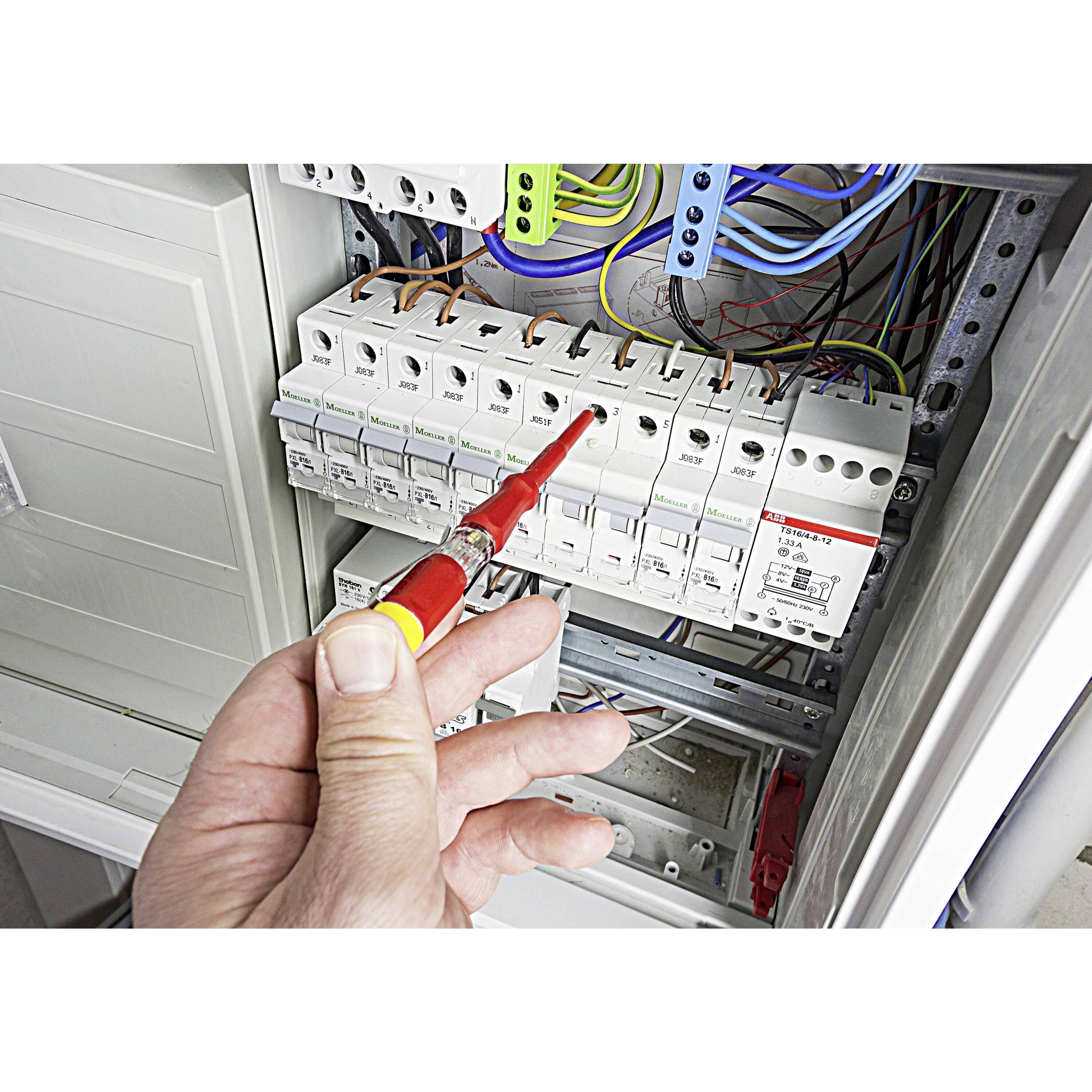 Une main tient un testeur de tension devant un boîtier électrique ouvert avec plusieurs fusibles et câbles disposés.