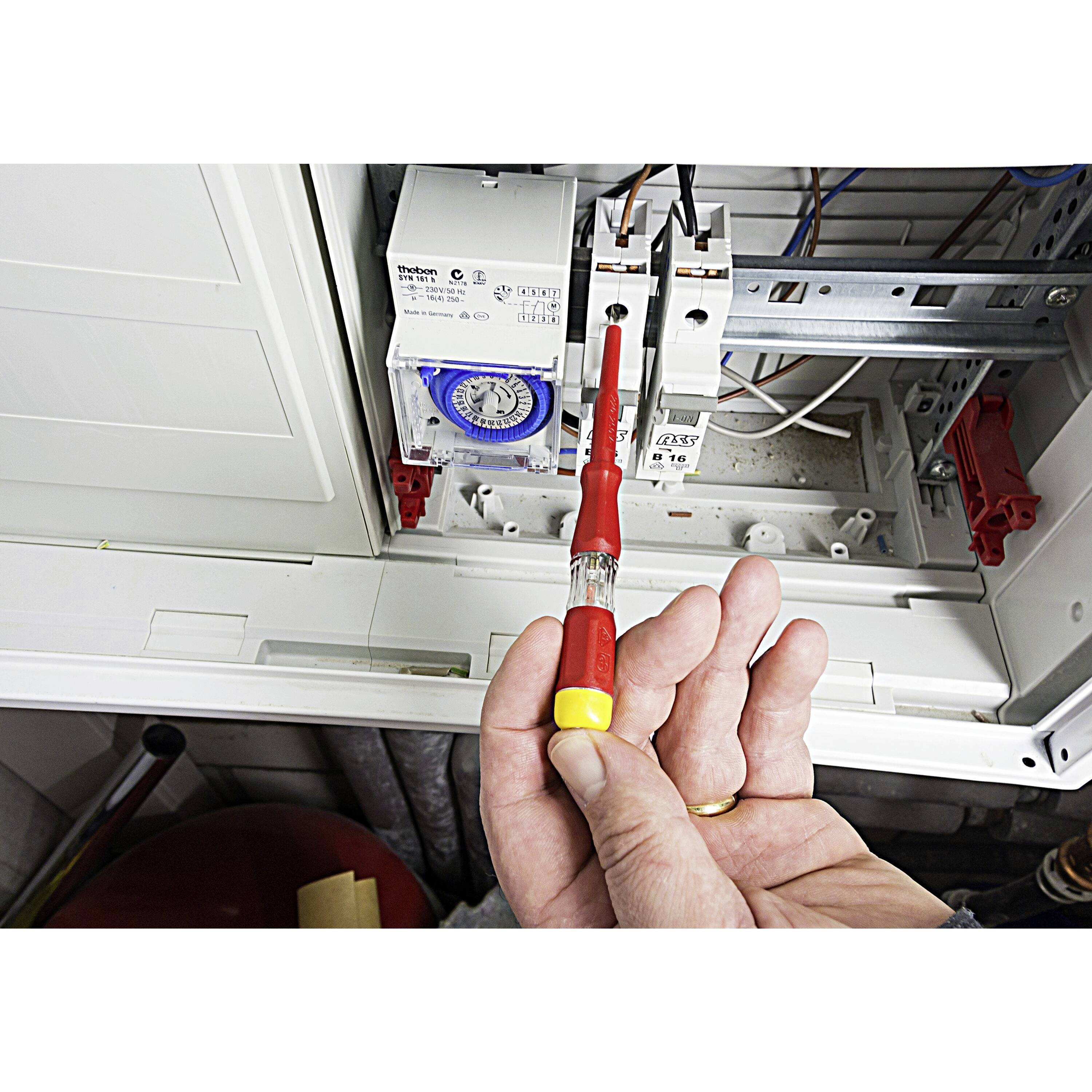 Une personne tient un testeur de tension devant un tableau électrique ouvert. Des fils et des interrupteurs sont visibles à l'intérieur.