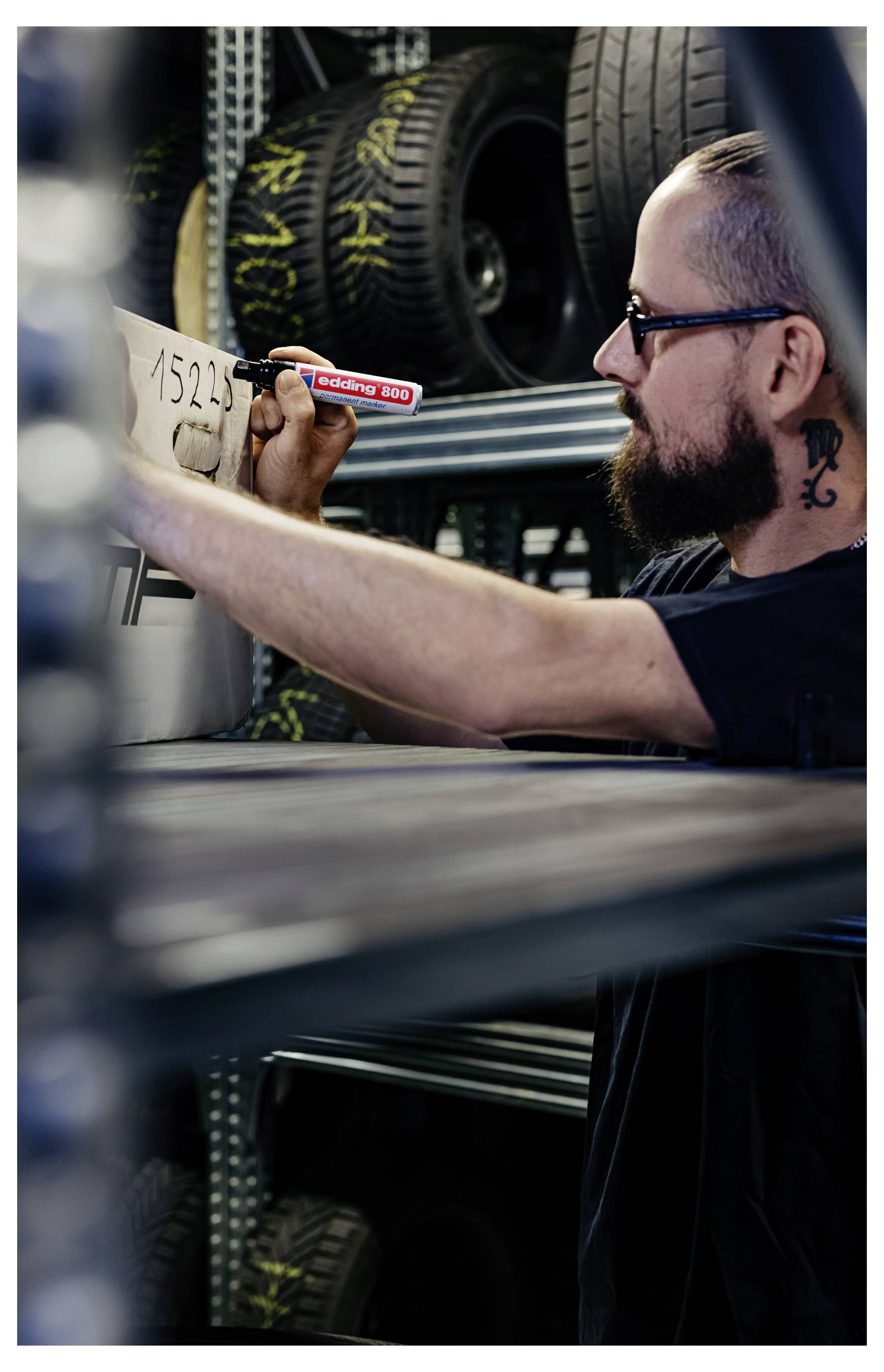 Homme barbu portant des lunettes écrivant sur le flanc d'un pneu avec un marqueur rouge dans un atelier, avec des pneus empilés en arrière-plan.