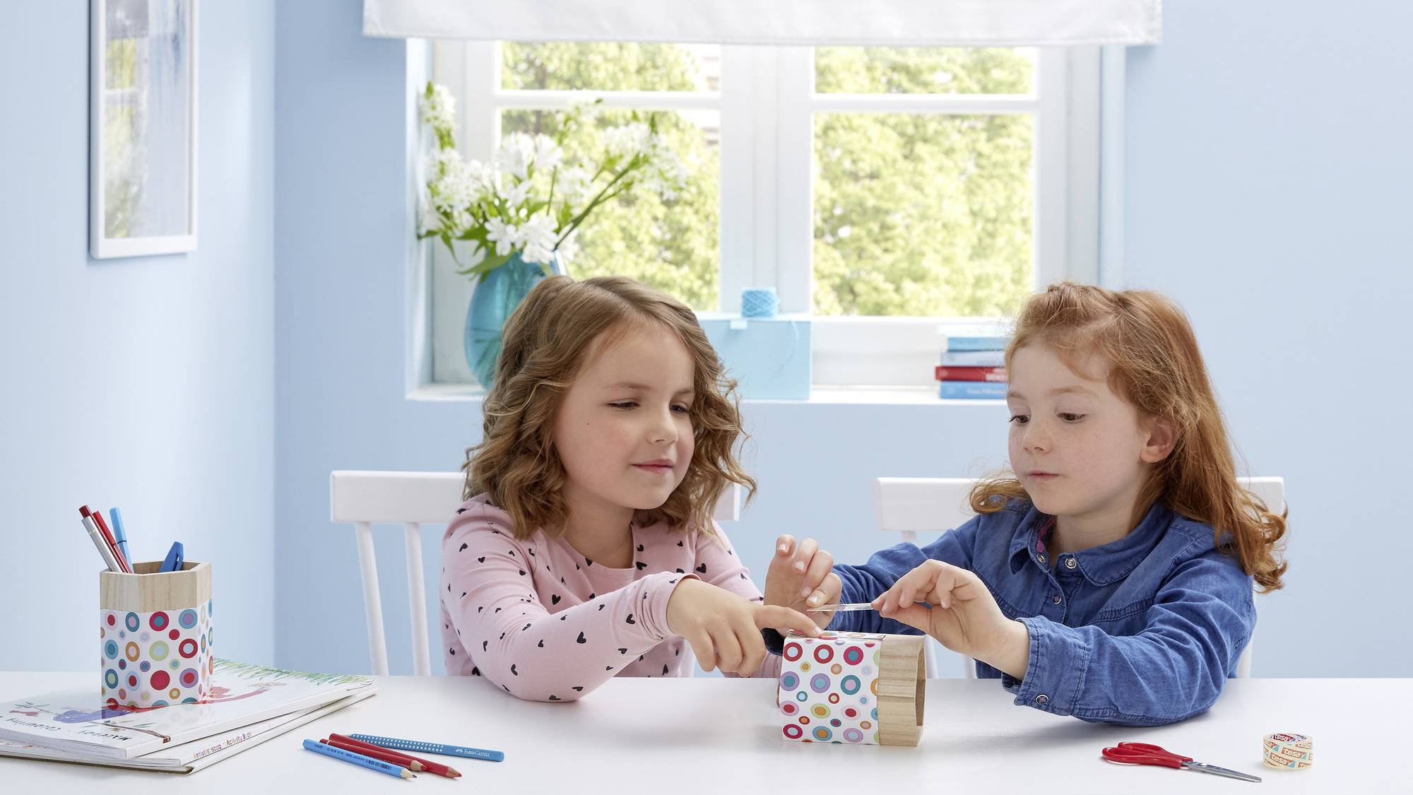 Deux enfants bricolent à une table dans une pièce lumineuse. Ils utilisent du papier coloré et des crayons. À l'arrière-plan, une fenêtre donne sur des plantes.