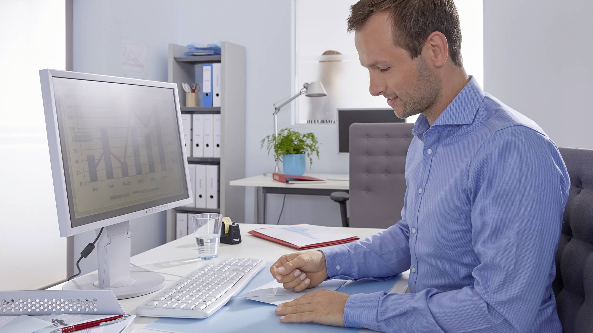 Un homme travaille dans un bureau sur des documents à son bureau, à côté d'un écran d'ordinateur affichant des diagrammes. Des étagères sont visibles en arrière-plan.