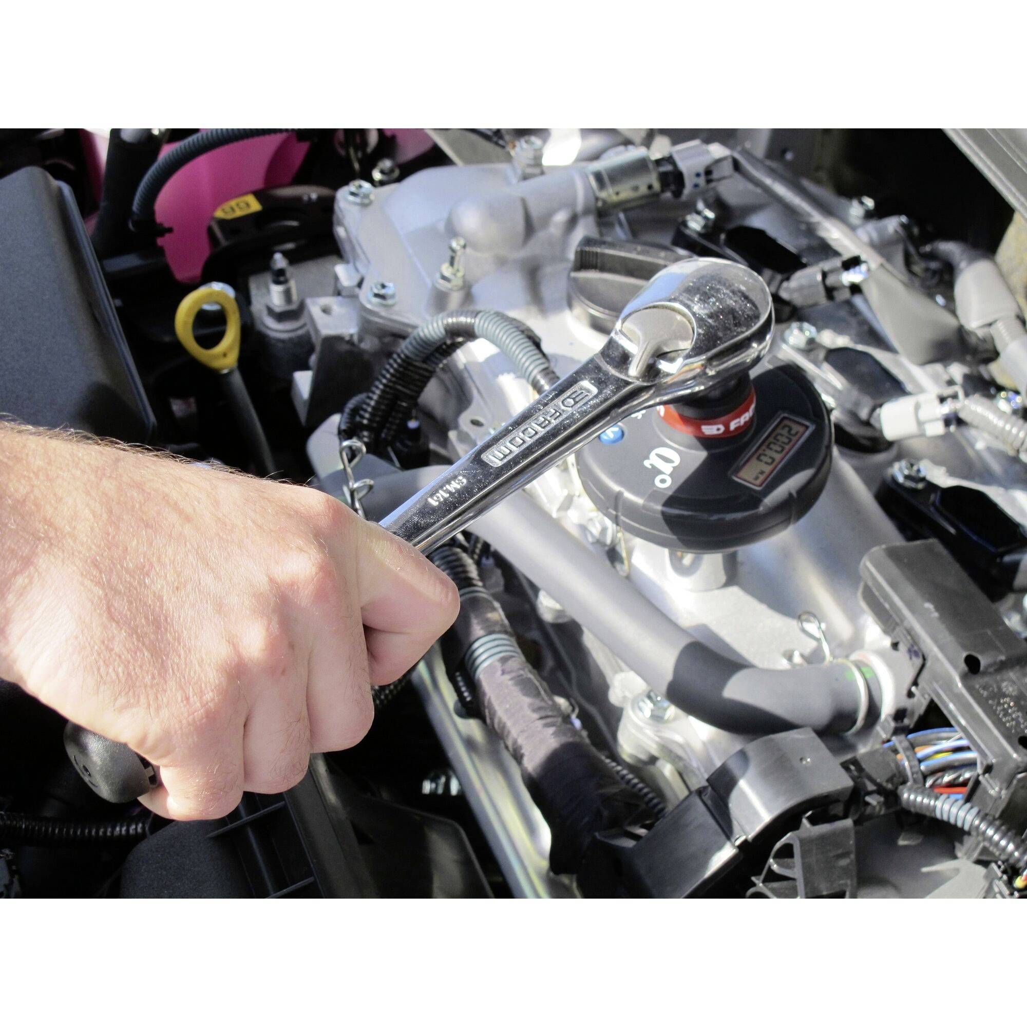 Une main tient une clé à molette et travaille sur un moteur de voiture, révélant de nombreux câbles et pièces mécaniques.