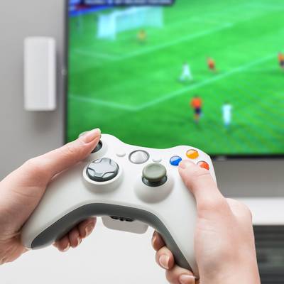 Une personne tient une manette de jeu vidéo alors qu'un match de football est affiché sur un écran. Les joueurs sont visibles en action.