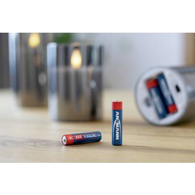 Deux piles AAA rouges et bleues sont posées sur une table ; en arrière-plan, un bougie brillante et une boîte de piles ouverte sont floues.