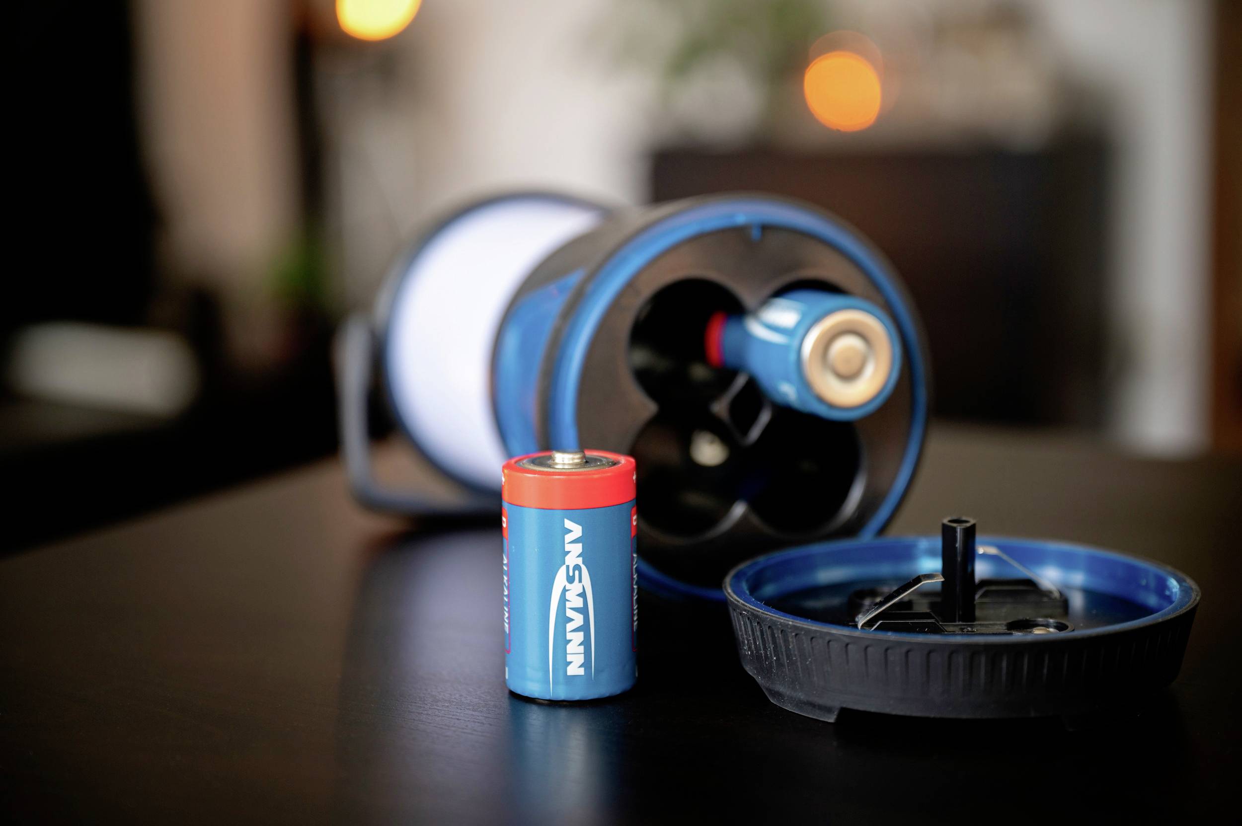 Une lampe de poche bleue démontée est posée sur une table, à côté d'une batterie unique. Des lumières floues sont visibles en arrière-plan.