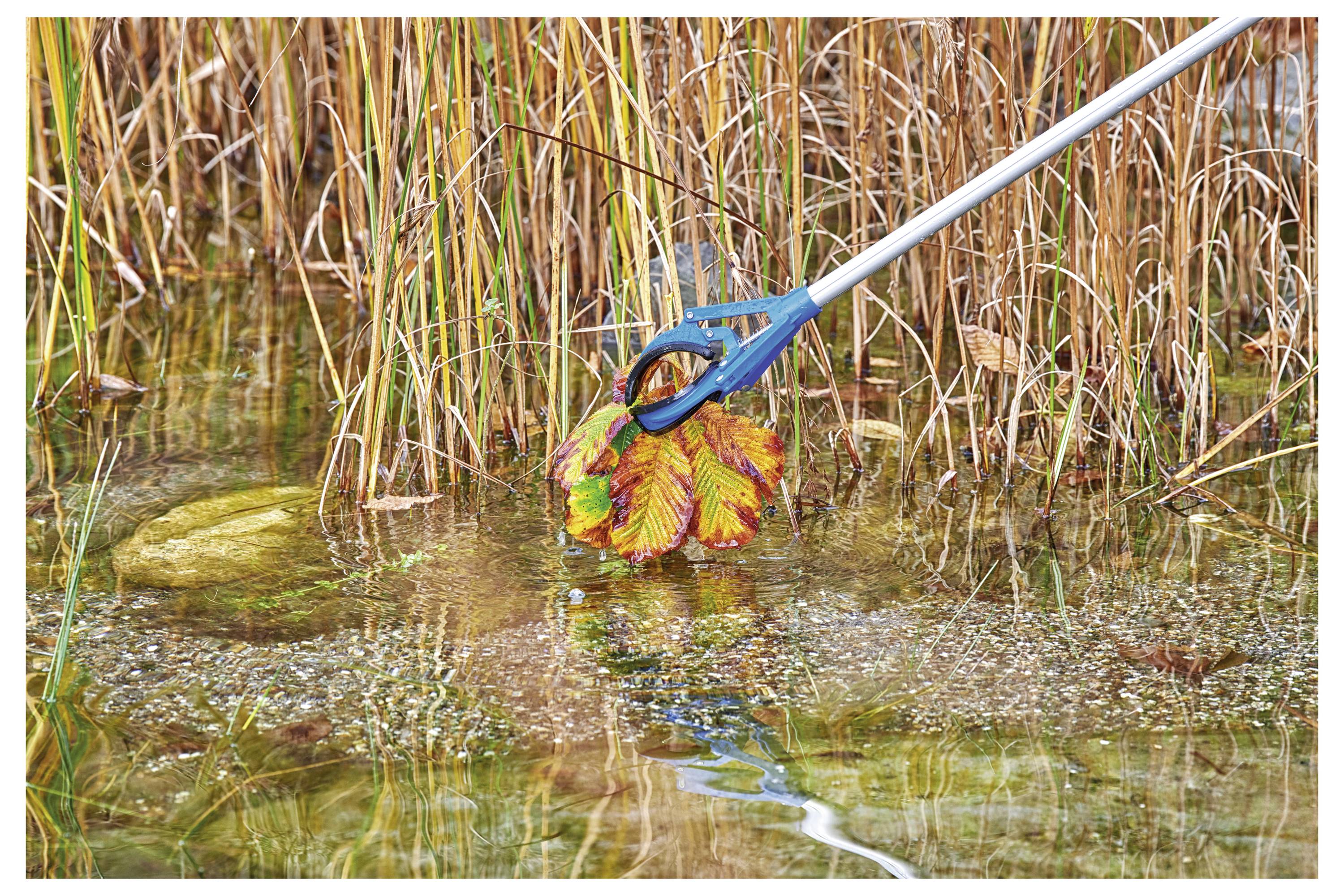 Un outil doté d'un manche bleu et d'une pince ramasse un objet en plastique coloré dans un étang, entouré de roseaux, lors d'un effort de nettoyage des eaux.