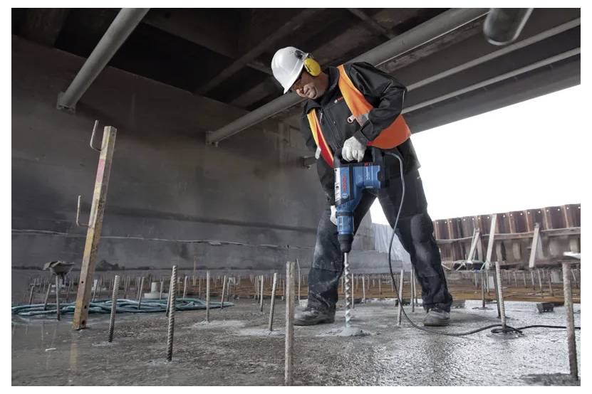 Un ouvrier du bâtiment portant un gilet de sécurité et un casque utilise un outil électrique pour percer du béton sur un chantier de construction, entouré de barres métalliques.