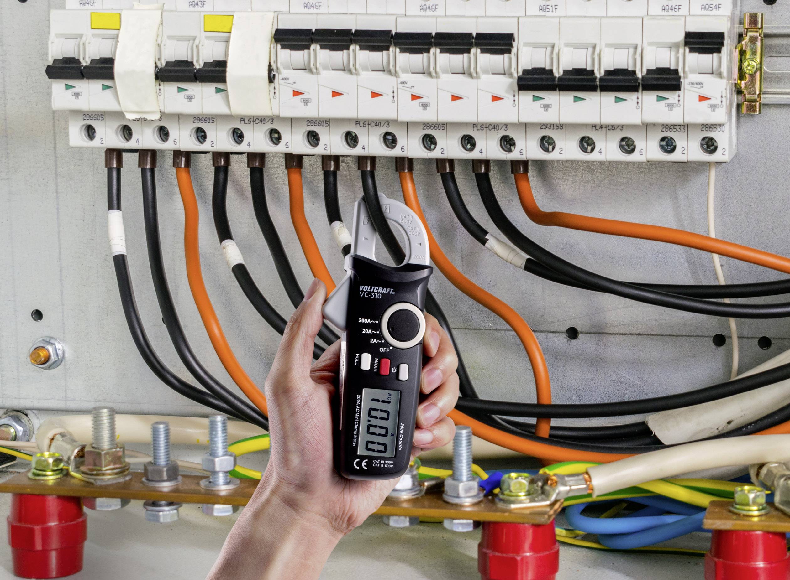 Une main tient un appareil de mesure électrique noir devant un tableau électrique ouvert comportant plusieurs câbles et disjoncteurs.