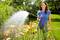 Une femme portant une chemise bleue arrose des lys oranges dans un jardin par une journée ensoleillée.