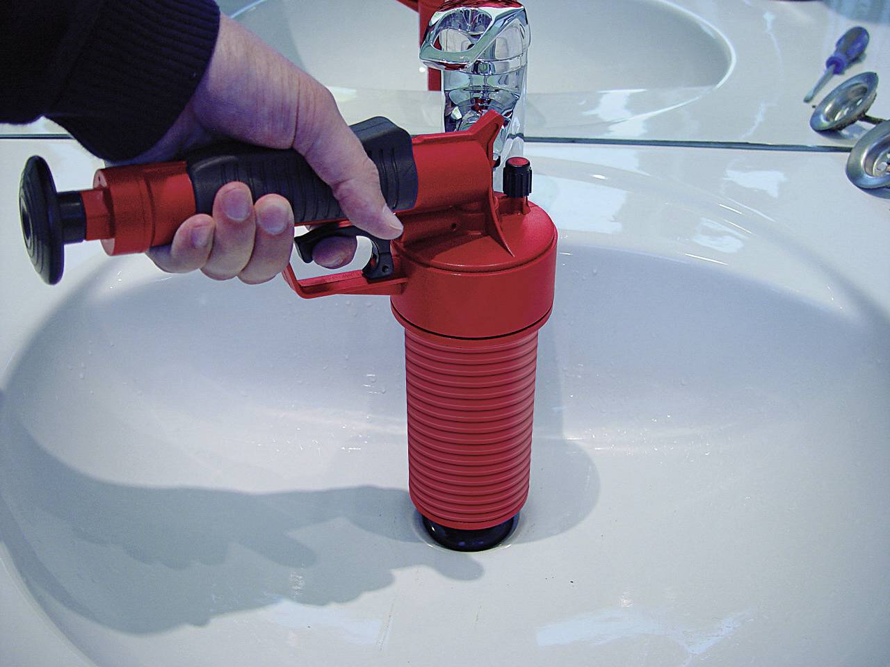 Une main tient un déboucheur rouge positionné au-dessus de l'évacuation d'un lavabo.