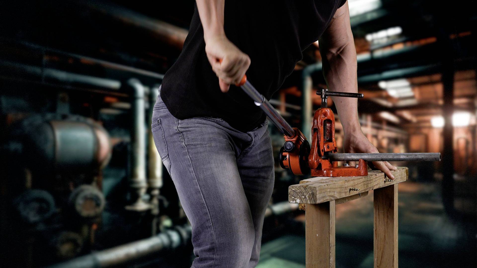 Une personne portant un t-shirt noir et un jean actionne avec force une machine à cintrer les tubes rouges dans un environnement industriel.