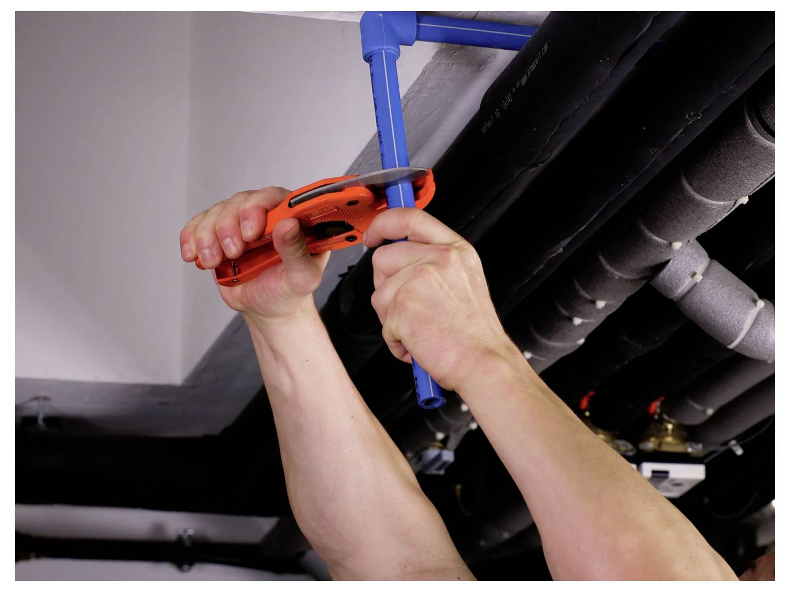 Les mains utilisent un coupe-tube orange pour couper un tuyau en plastique bleu sous la tuyauterie au plafond.