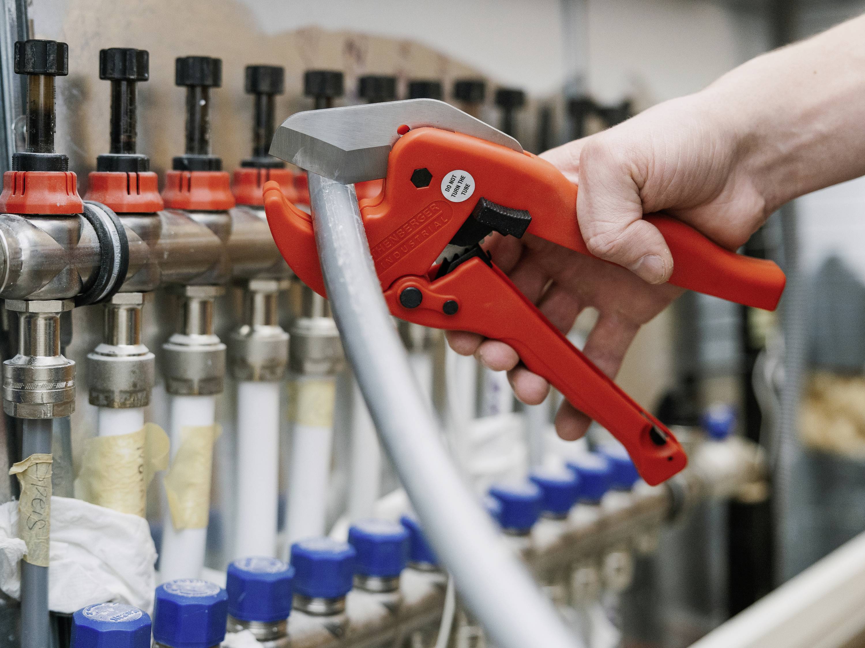 Une personne coupe un tuyau gris avec un coupe-tube rouge à côté de plusieurs conduites et vannes.