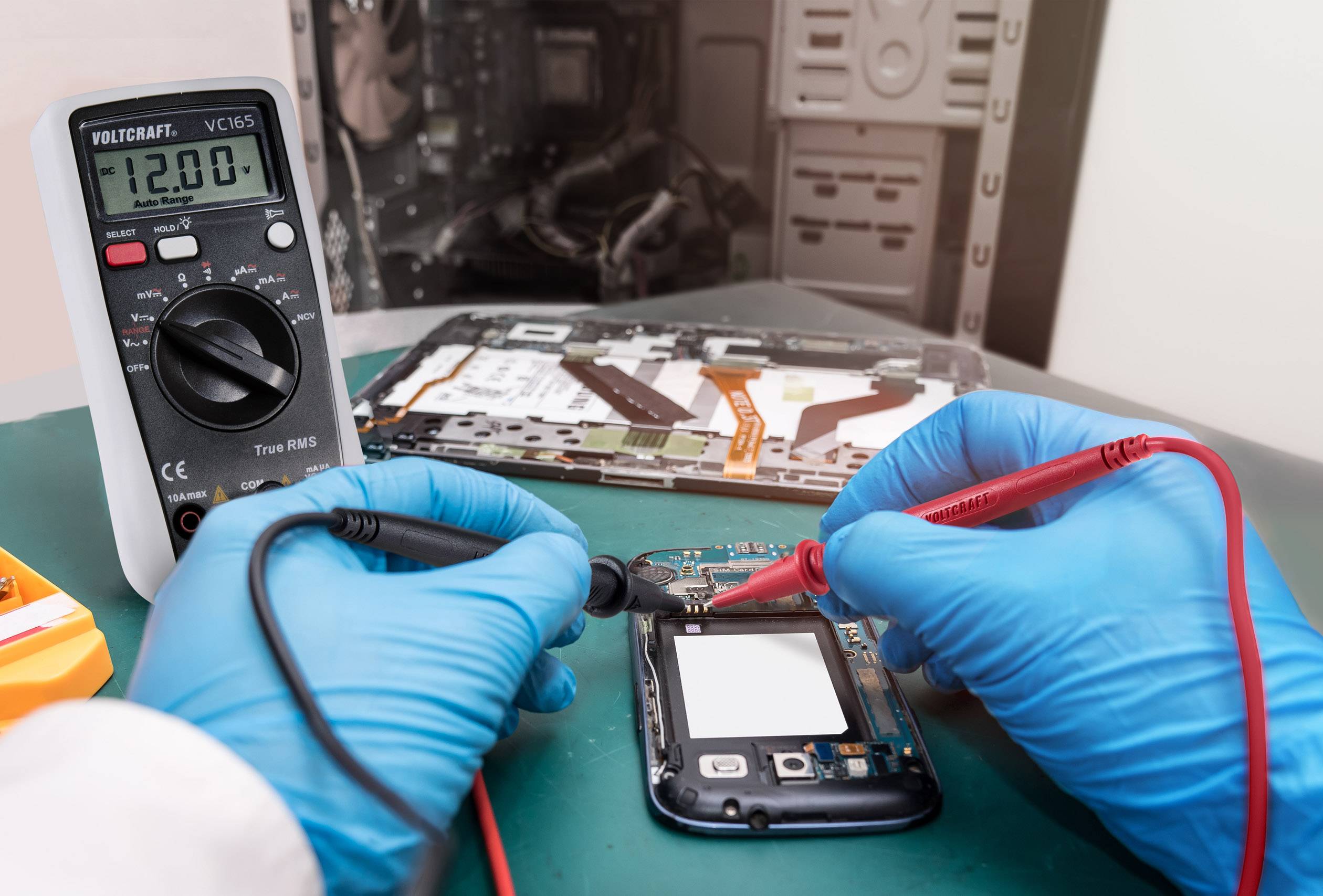 Un technicien répare un smartphone. Des gants bleus tiennent des sondes de test contre l'appareil, un multimètre affiche '12,0' volts. Un ordinateur ouvert est visible en arrière-plan.