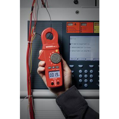 Une personne tient un multimètre numérique rouge devant un armoire électrique et mesure la tension ; l'affichage indique '00.19'.