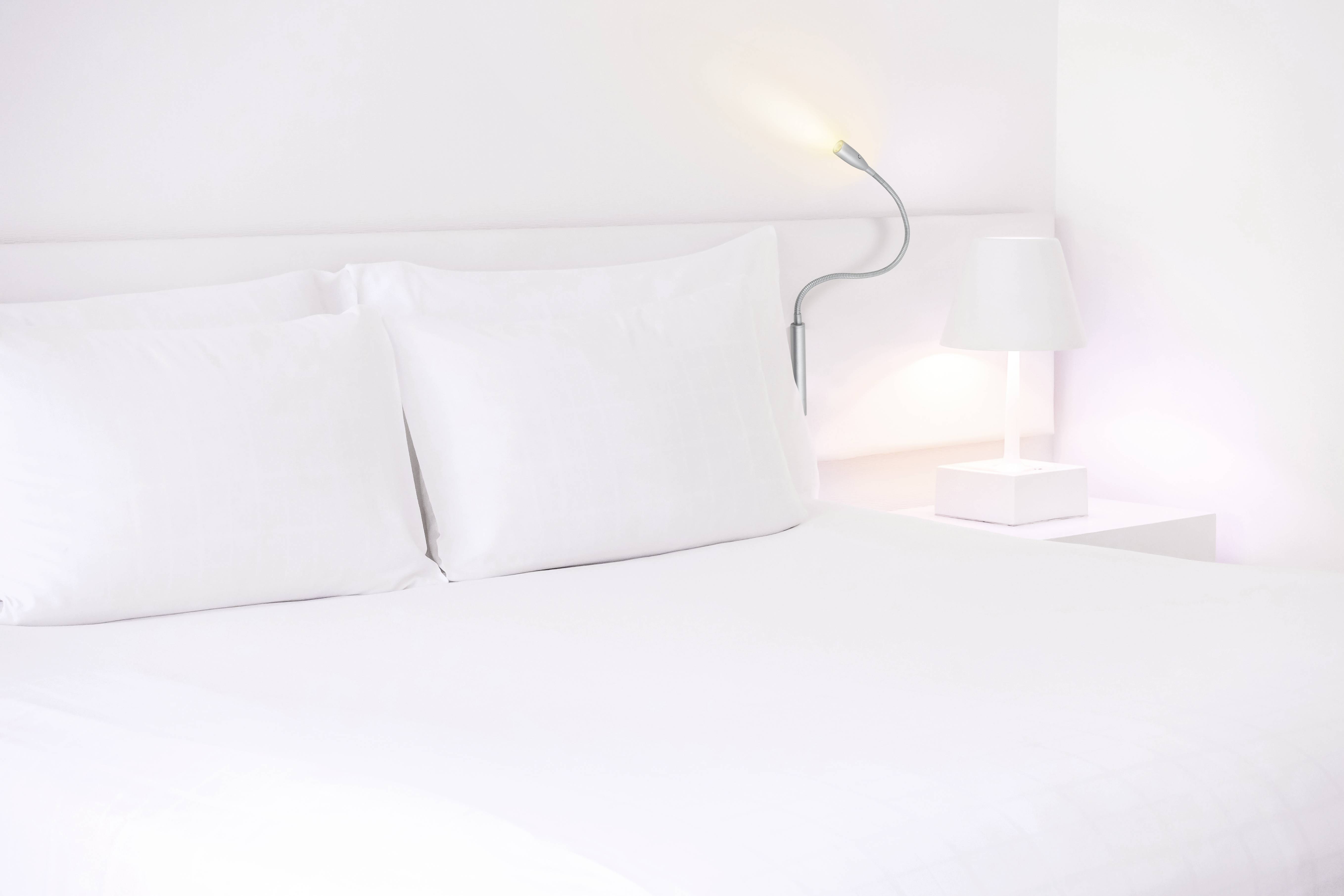 Un lit blanc avec deux oreillers se trouve dans une pièce lumineuse et minimaliste. Une lampe et une table de chevet sont à côté.