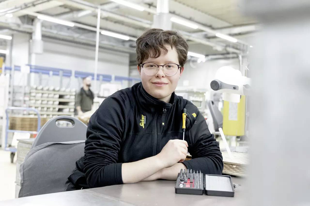 Une personne souriante portant des lunettes est assise dans un atelier. Elle porte un pull noir et tient plusieurs outils. Des étagères et une autre personne sont visibles en arrière-plan.