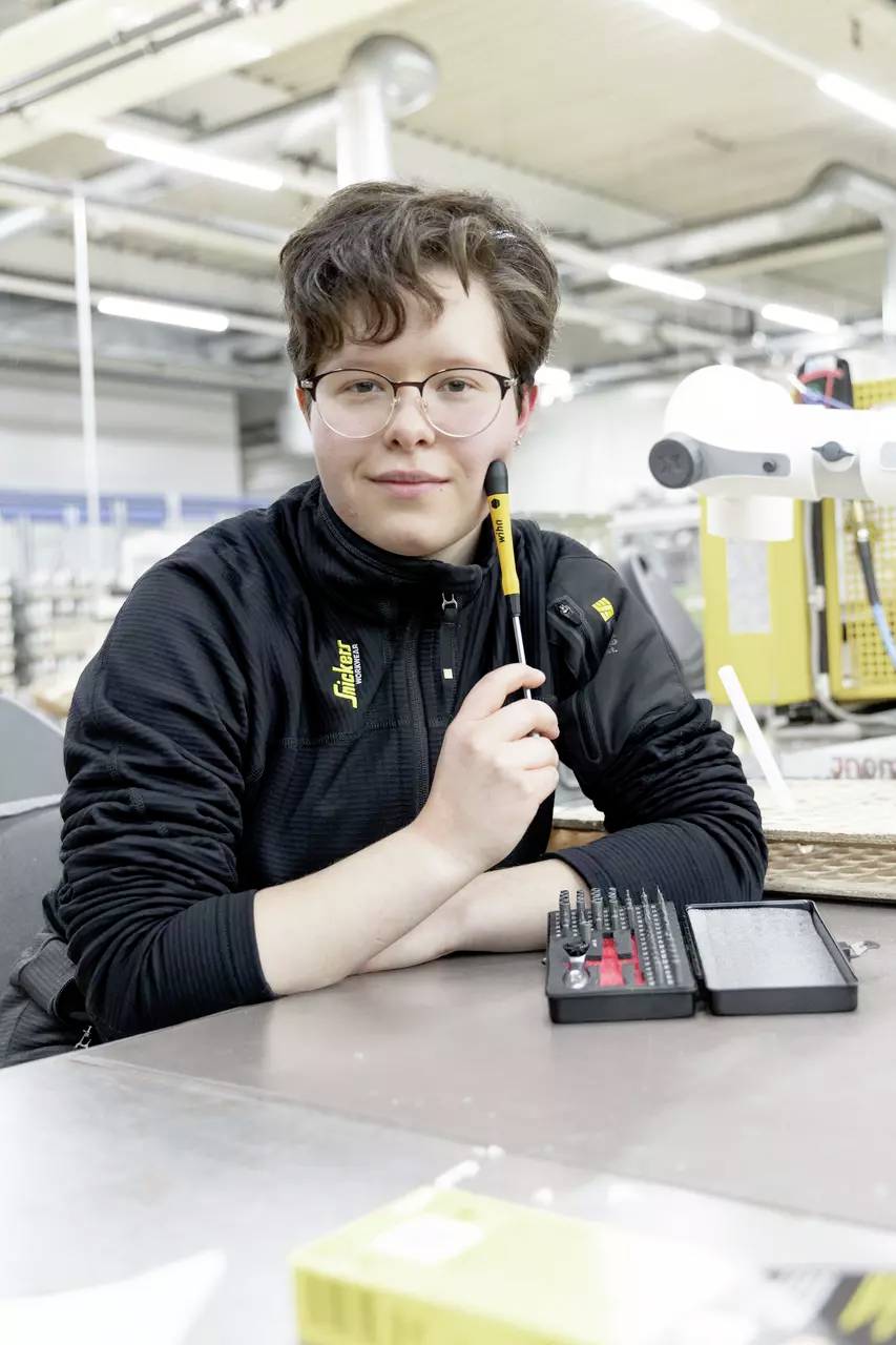 Une personne dans un atelier tient un stylo et sourit à la caméra. Des outils sont posés sur la table. Des équipements industriels sont visibles en arrière-plan.
