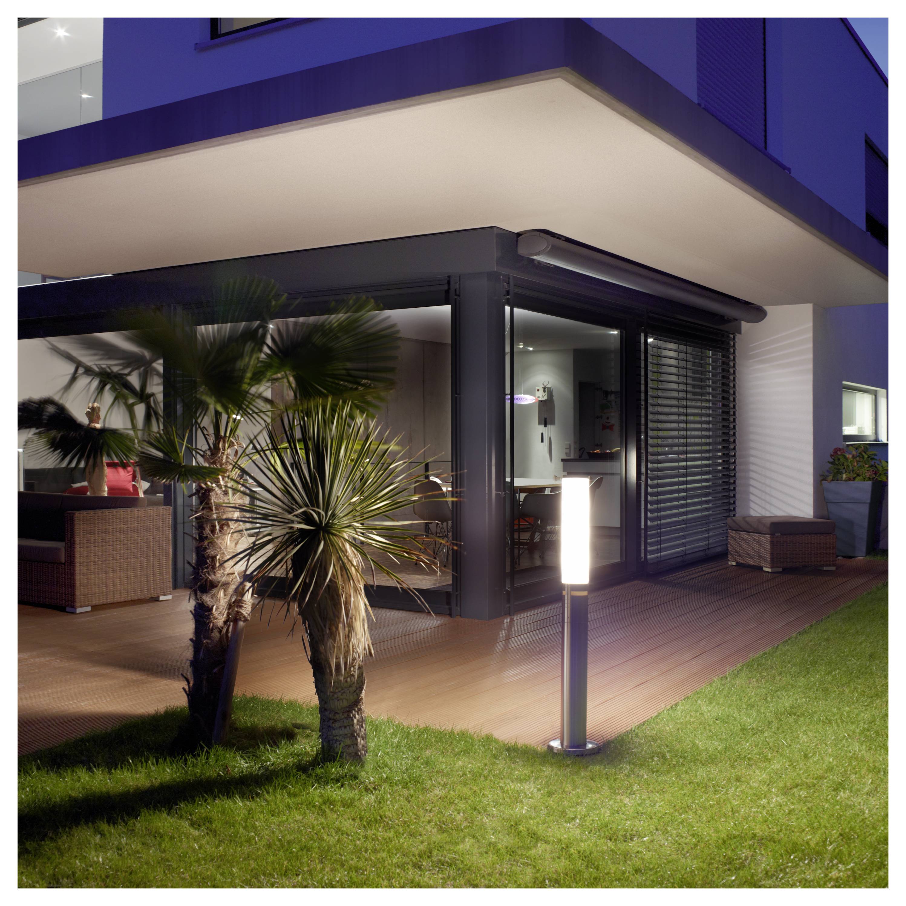 Patio de maison moderne au crépuscule avec poteau de jardin illuminé, plantes de palmier, terrasse en bois et grandes portes en verre sous un toit en porte-à-faux.