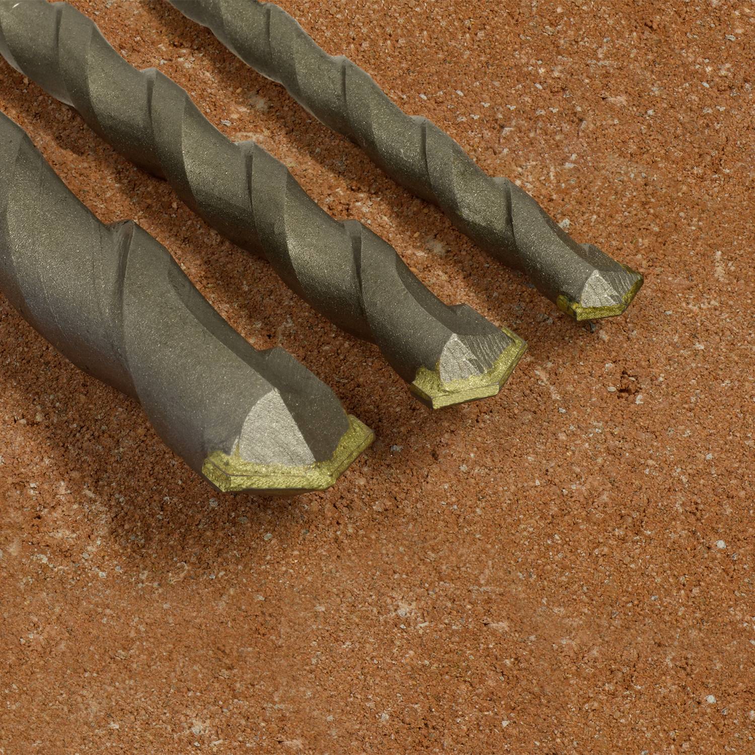 Trois forets de tailles différentes, au design hélicoïdal et aux pointes métalliques, sont posés sur une surface rugueuse de couleur rougeâtre.