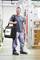 Un homme se tient dans un atelier et porte une mallette à outils noire avec le logo 'wiha'. Il porte un t-shirt gris et un pantalon de travail.
