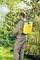 Une personne vêtue de tenue de travail beige pulvérise des plantes en plein air avec un pulvérisateur à dos jaune. Elle se trouve dans un jardin luxuriant.