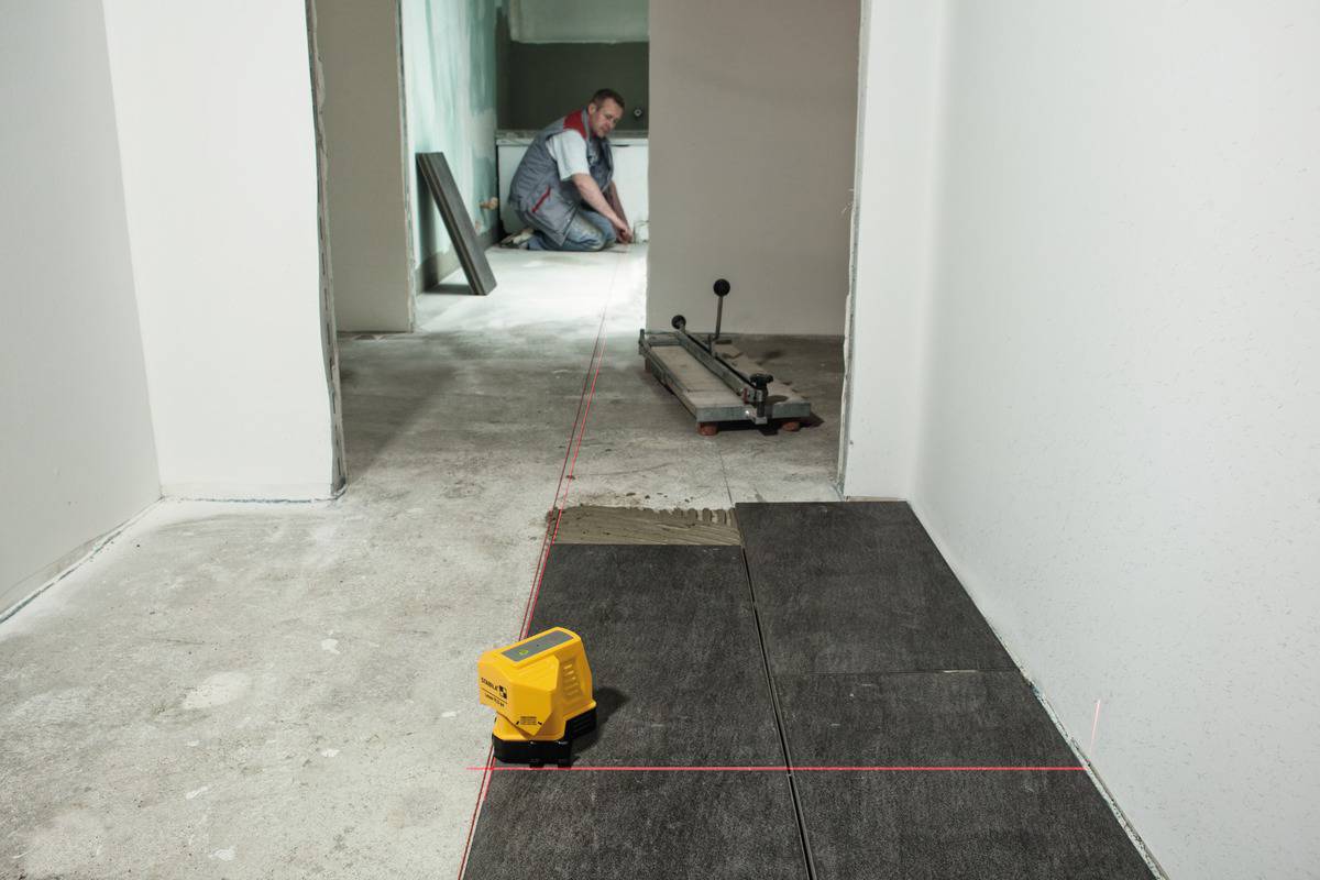 Un artisan pose des carreaux de sol dans une pièce. Un appareil de nivellement laser projette des lignes rouges pour l'alignement.