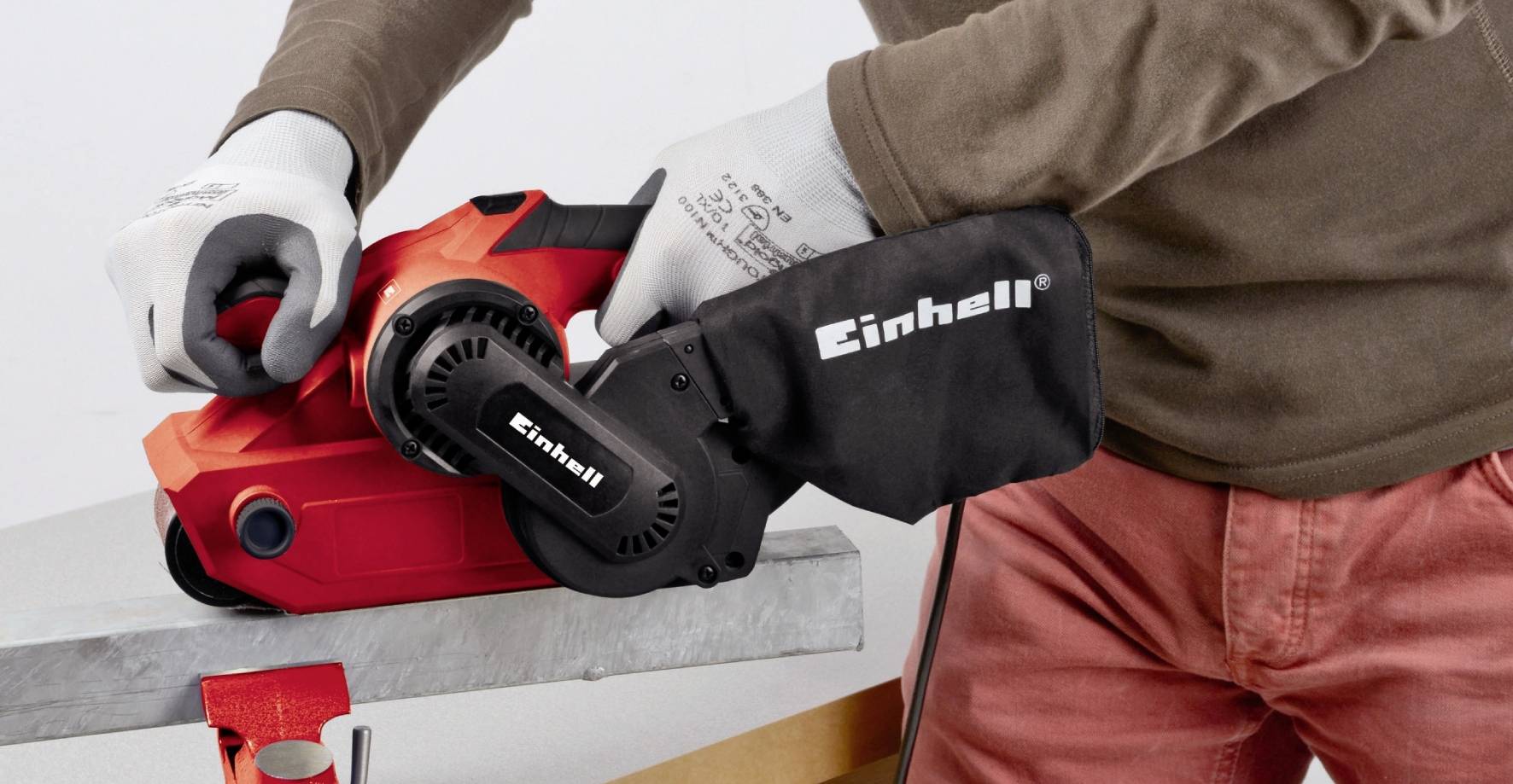 Une personne ponçe une surface métallique avec une ponceuse à bande rouge. La ponceuse est équipée d'un sac à poussière noir portant le logo 'Einhell'.