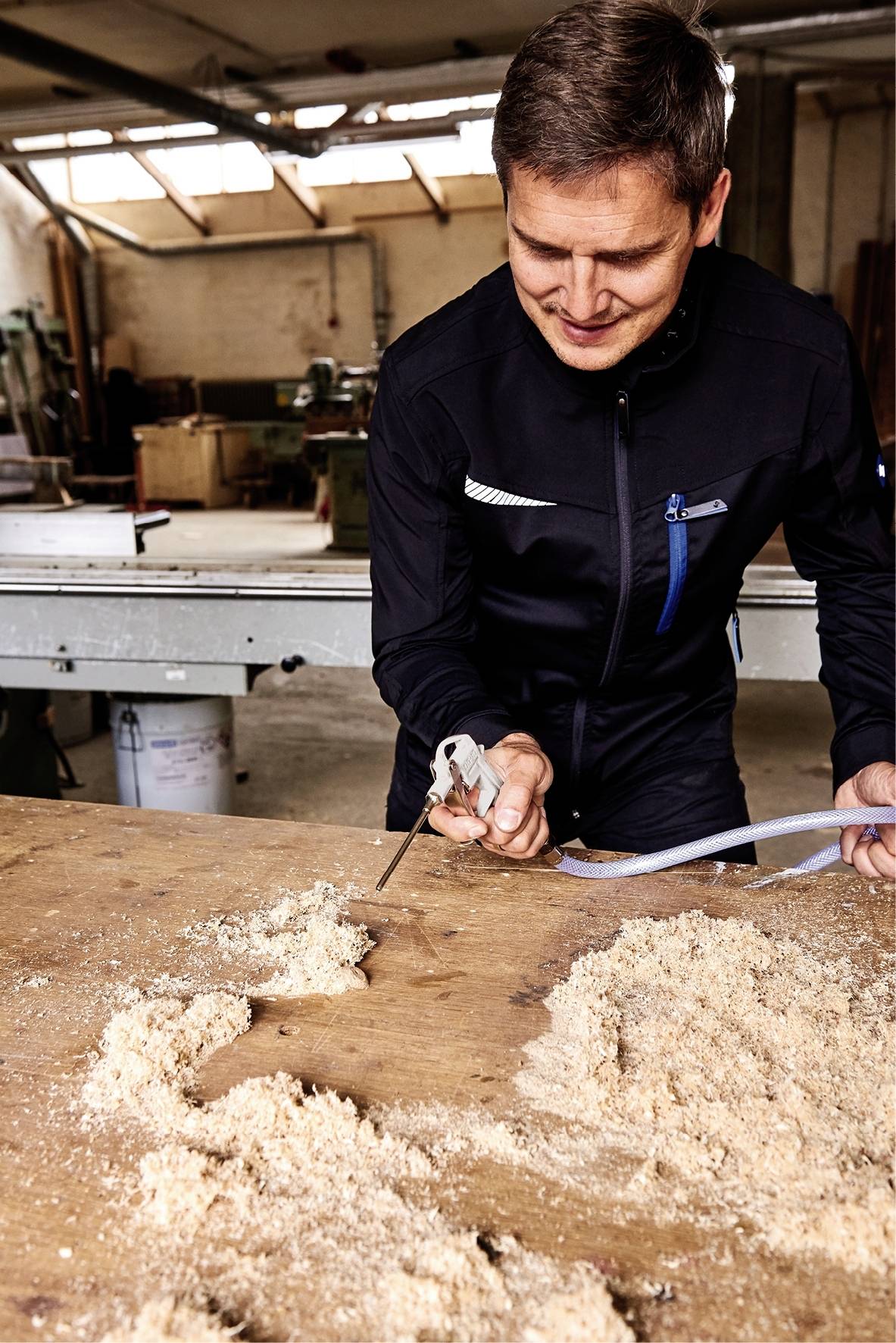 Un homme coupe un morceau de matériau avec un outil dans un atelier, sur une table couverte de sciure.