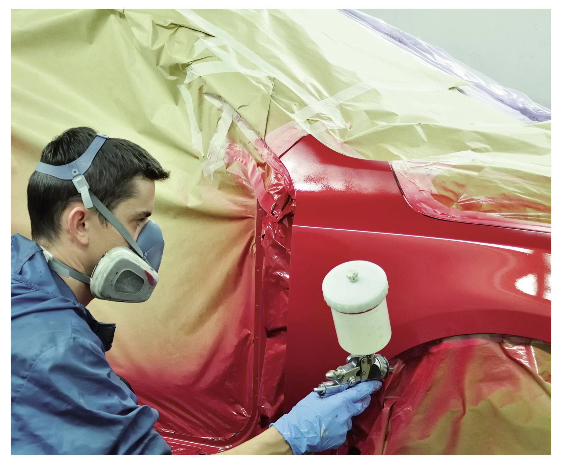 Un technicien en masque et gants peint au pistolet un panneau de voiture rouge dans un atelier de carrosserie, le reste du véhicule étant protégé.